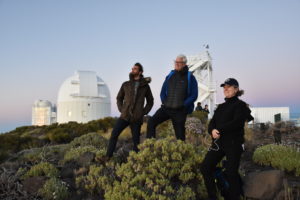 two men and a woman stand on a hill looking off into the distance with a white observatory behind them
